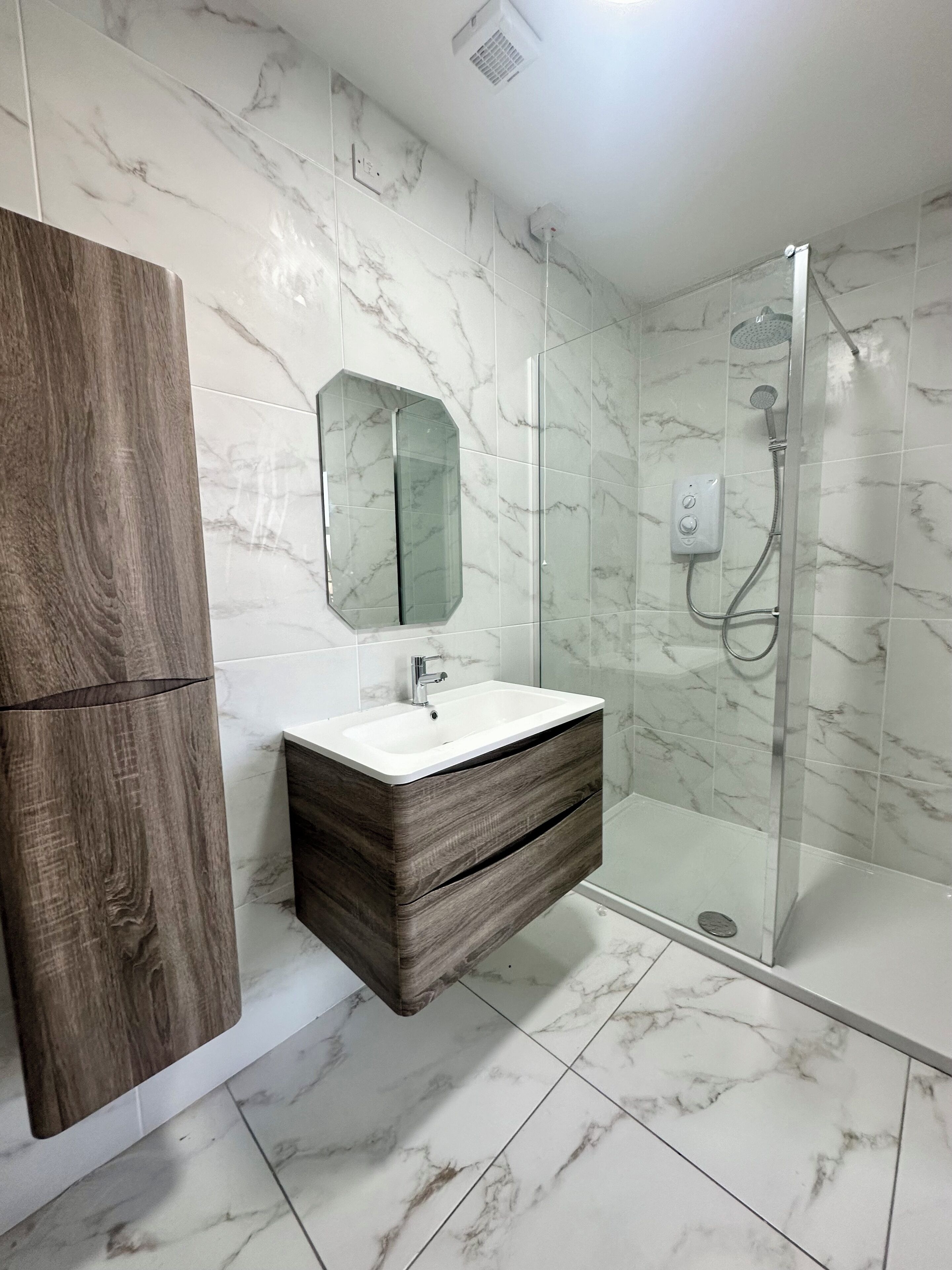 Un baño moderno con ducha de azulejos de mármol, tocador de madera con espejo y lavamanos blanco.