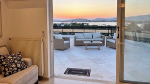 Un balcón con sofá y sillas, vista a una puesta de sol sobre un paisaje urbano, y un radiador en la habitación.