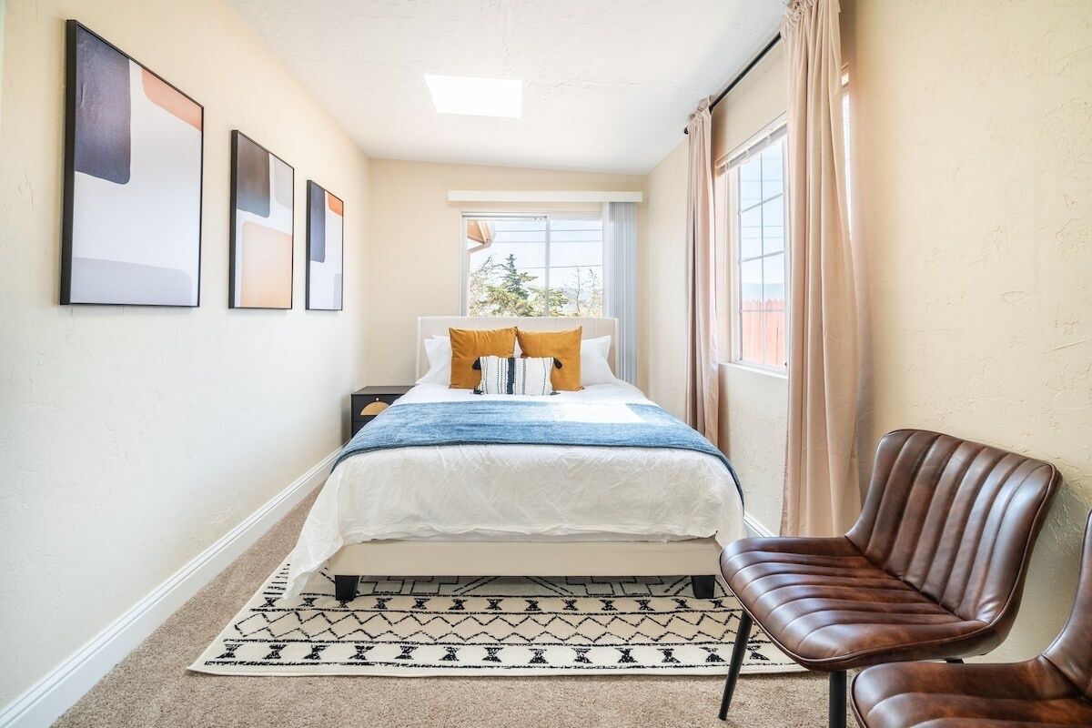 A bedroom with a bed, a chair, a window, and framed artwork on the wall.