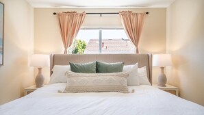 A neatly made bed with white linens and decorative pillows, two bedside lamps, and a window with curtains.