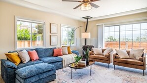 A living room with a blue sofa, a brown sofa, and a coffee table. There are large windows and a ceiling fan.