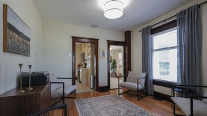 Uma sala de estar moderna com piso de madeira, um tapete estampado, um sofá, uma poltrona, uma mesinha de apoio com toca-discos e uma janela grande com cortinas.