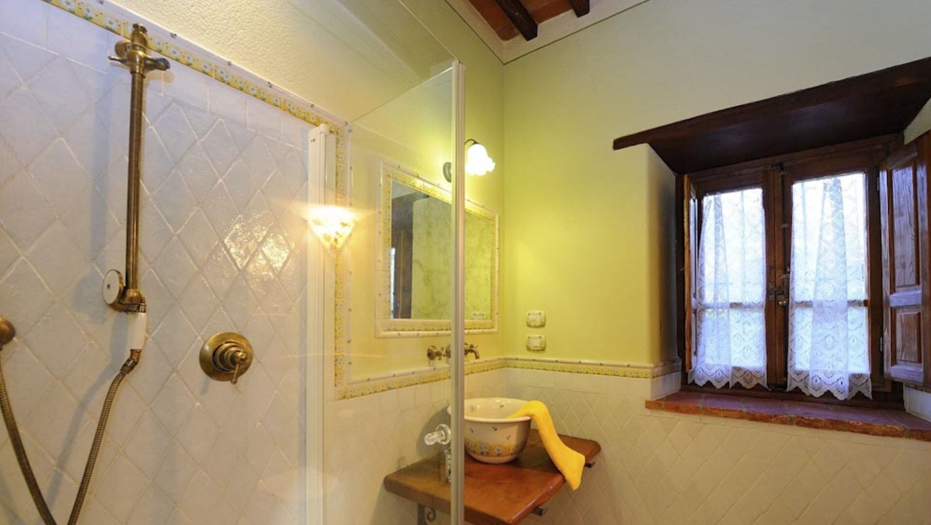 Un baño con ducha, lavamanos, espejo y ventana con cortinas de encaje.