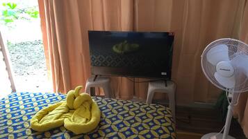 Une chambre avec un lit, un téléviseur posé sur un meuble et un ventilateur sur pied.