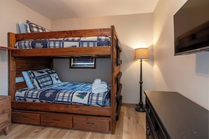 Una cama litera con colcha, una lámpara de noche y un televisor en la pared.