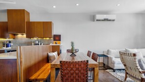 A modern kitchen with wooden cabinets, a dining area with a wooden table and chairs, and a living area with a sofa and patterned chairs.