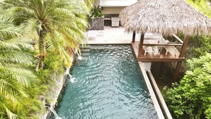 A pool with a thatched roof structure, surrounded by lush greenery and palm trees.