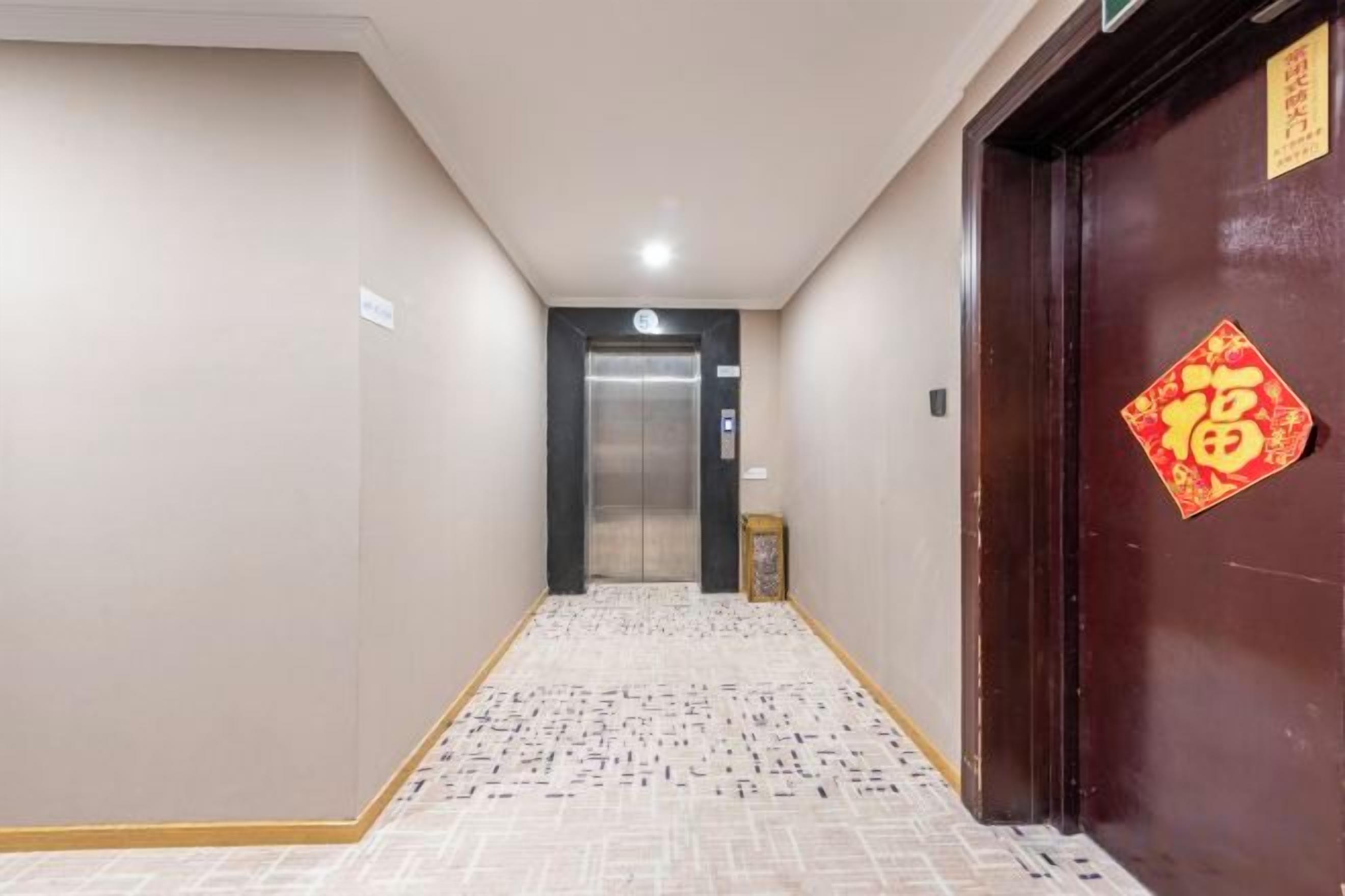 Un couloir avec un sol à motifs, une porte ornée d’un décor rouge et un ascenseur visible au loin.