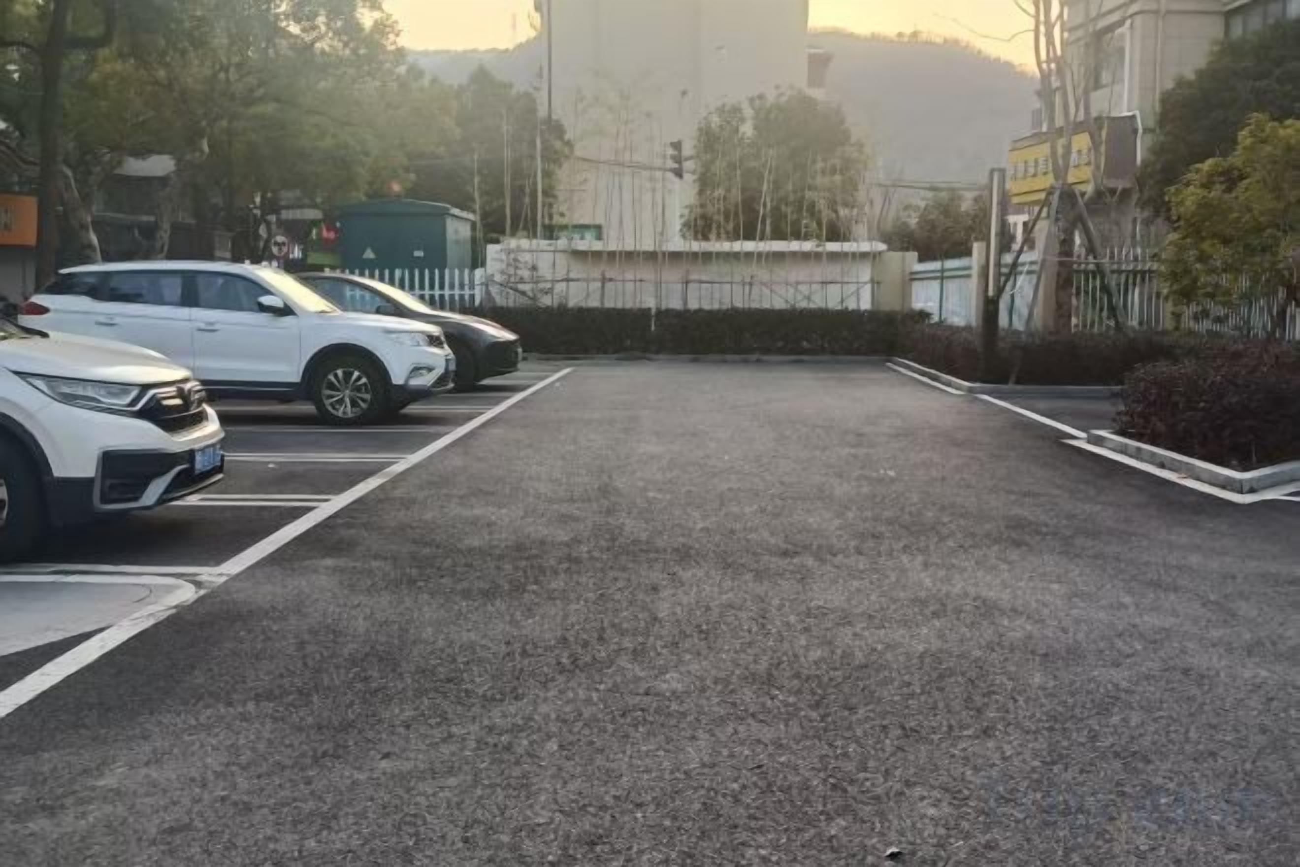 Een parkeerplaats met diverse auto's, een hek en gebouwen op de achtergrond.