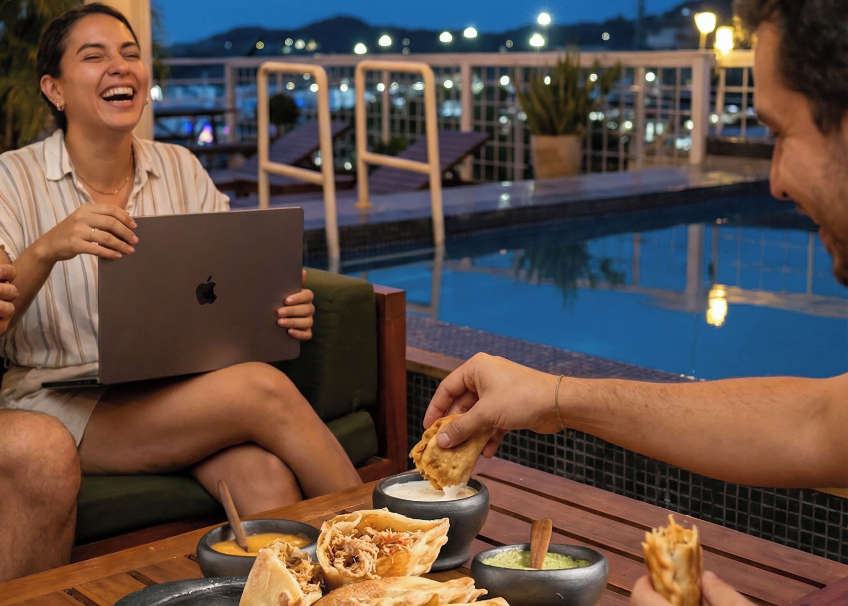 Two people seated by a poolside table with food and drinks, a laptop, and a pool in the background.