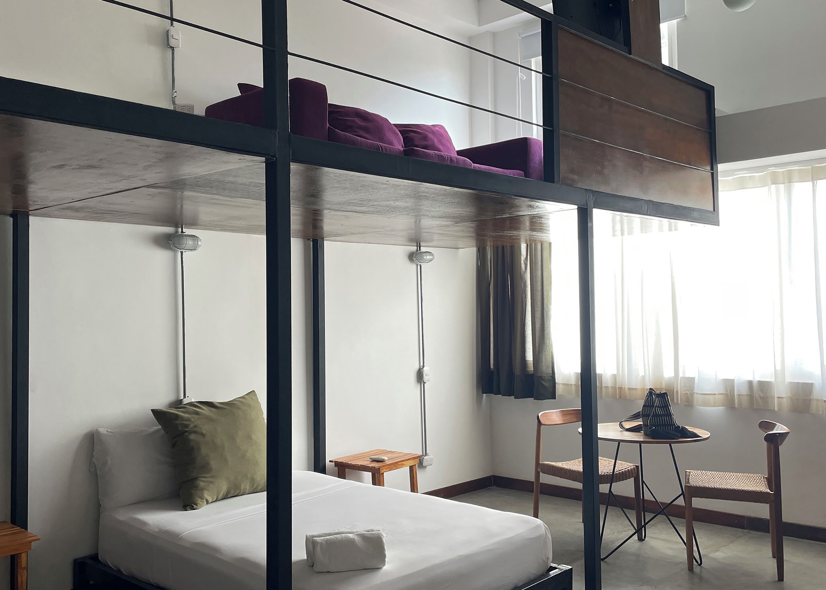 Una cama individual superpuesta con un sofá cama debajo, una pequeña mesa de madera, una silla y una ventana con cortinas.
