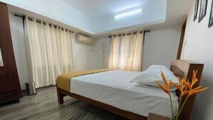 A hotel room with a bed, curtains, a wooden headboard, a bedside table with a vase of flowers, and an air conditioning unit.