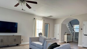 A living room with a flat-screen TV, two grey sofas, a ceiling fan, and a staircase leading to an upper level.