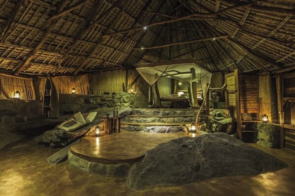A rustic interior with a thatched roof, stone walls, and a central stone platform with seating.