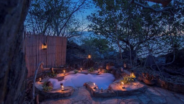 Un espace extérieur paisible avec un jacuzzi, agrémenté de sièges en pierre et de bougies, entouré d’arbres et d’une clôture en bois.