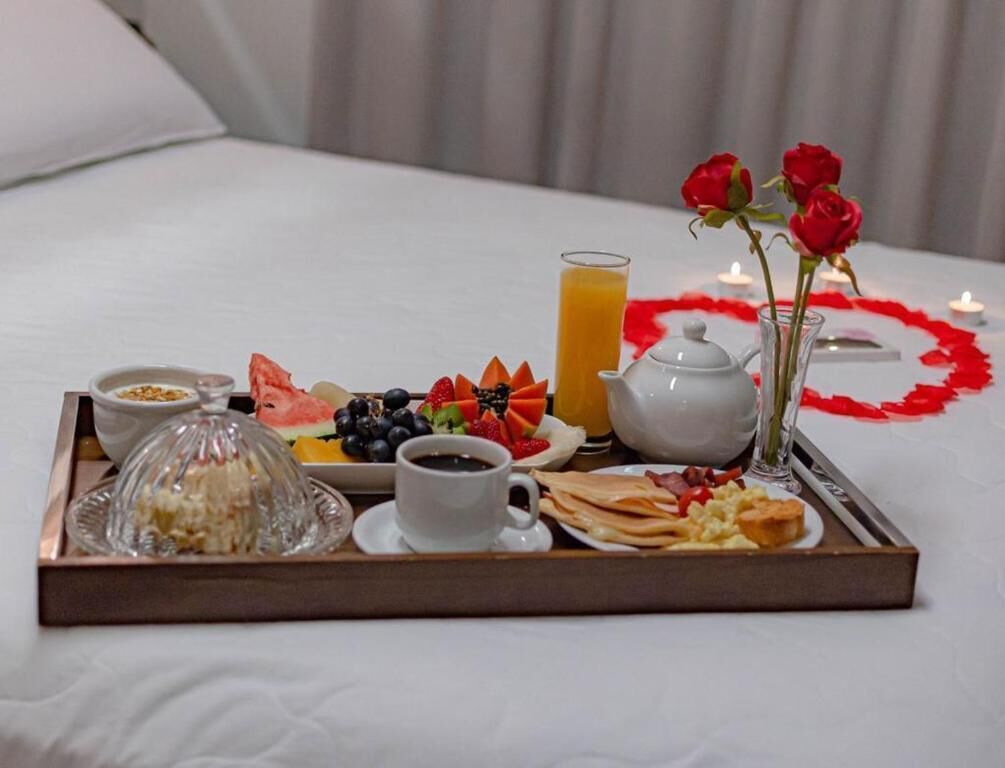 Una bandeja de desayuno sobre la cama con diversos alimentos, un vaso de zumo de naranja, una tetera y un florero con rosas.