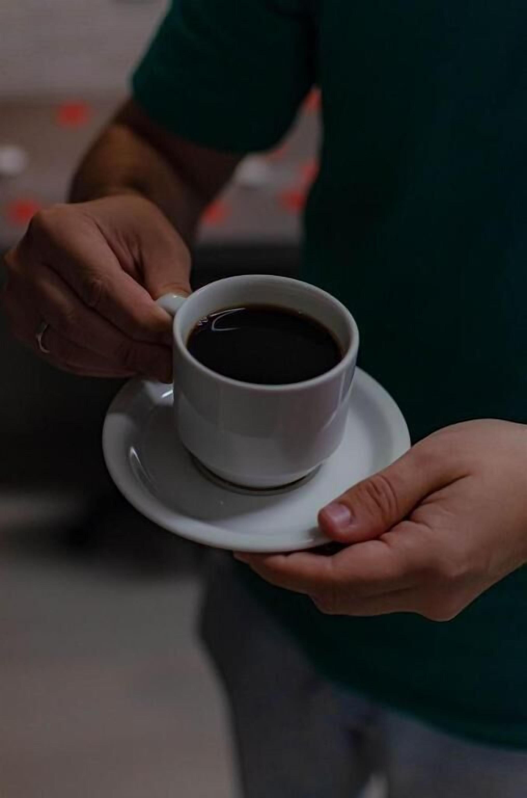 Eine Person hält eine weiße Tasse mit schwarzer Flüssigkeit, vermutlich Kaffee, auf einem Untertasse.