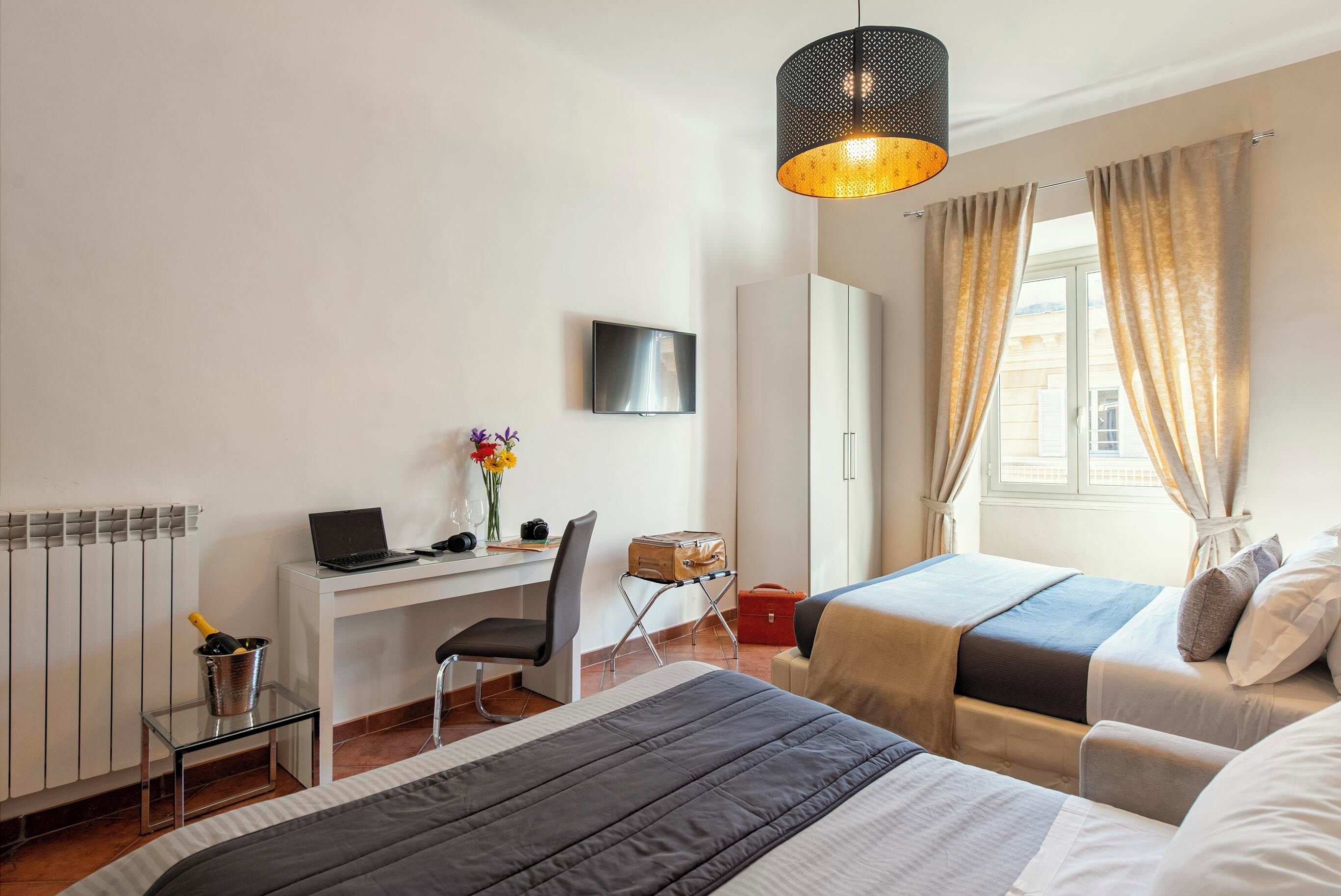 Ein Hotelzimmer mit einem Bett, einem Schreibtisch mit Laptop, einem Sessel, einem Fernseher und einem Fenster mit Vorhängen.