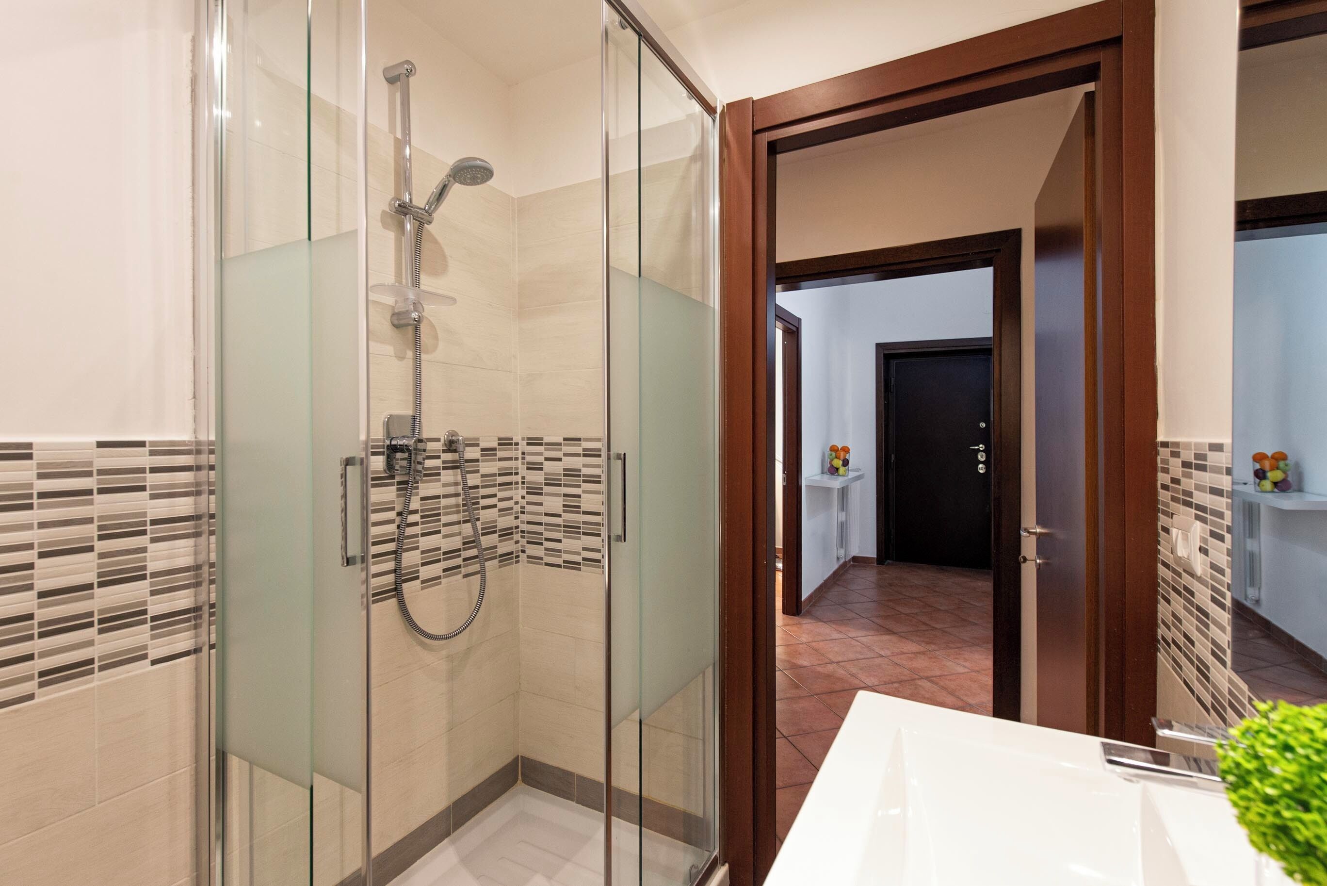 Une salle de bain moderne avec une douche à parois de verre, un lavabo blanc et une porte en bois donnant sur une autre pièce.