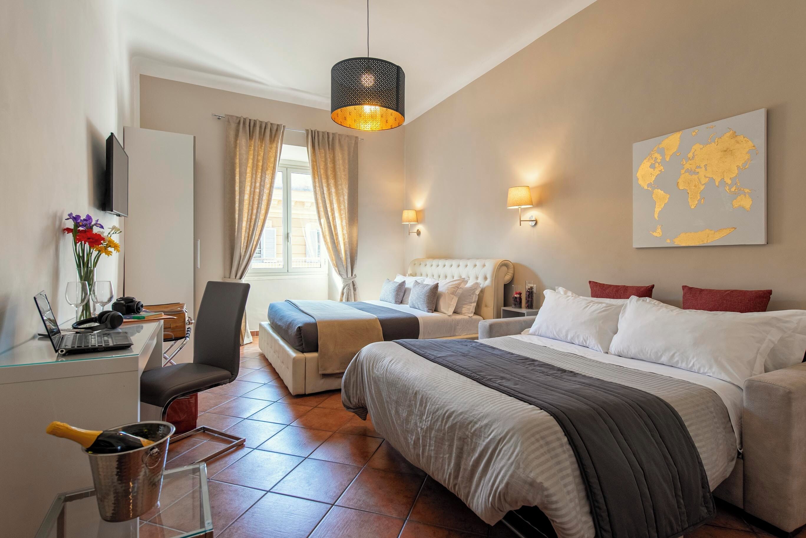Une chambre d’hôtel avec un lit, un bureau avec un ordinateur, une chaise, une petite table avec une boisson, et une carte du monde accrochée au mur.
