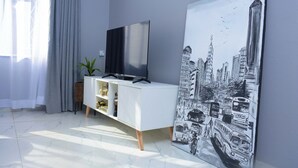 Una sala moderna con un televisor de pantalla plana sobre un mueble blanco, un cuadro de gran tamaño con un paisaje urbano y una planta en maceta.