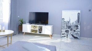 Una sala moderna con un televisor de pantalla plana sobre un mueble blanco, un cuadro de un paisaje urbano y una mesa redonda con velas.