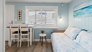 A room with a bed, a dining table with chairs, a small side table, and a window with blinds.