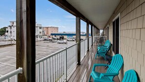 Een overdekt buitengebied met een houten terras, turquoise stoelen en een uitzicht op een parkeerplaats en gebouwen op de achtergrond.