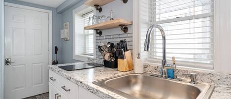 A kitchen with a granite countertop, stainless steel sink, wooden shelves, and a window with blinds.