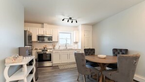 A modern kitchen with white cabinets, stainless steel appliances, and a dining area with a wooden table and grey upholstered chairs.