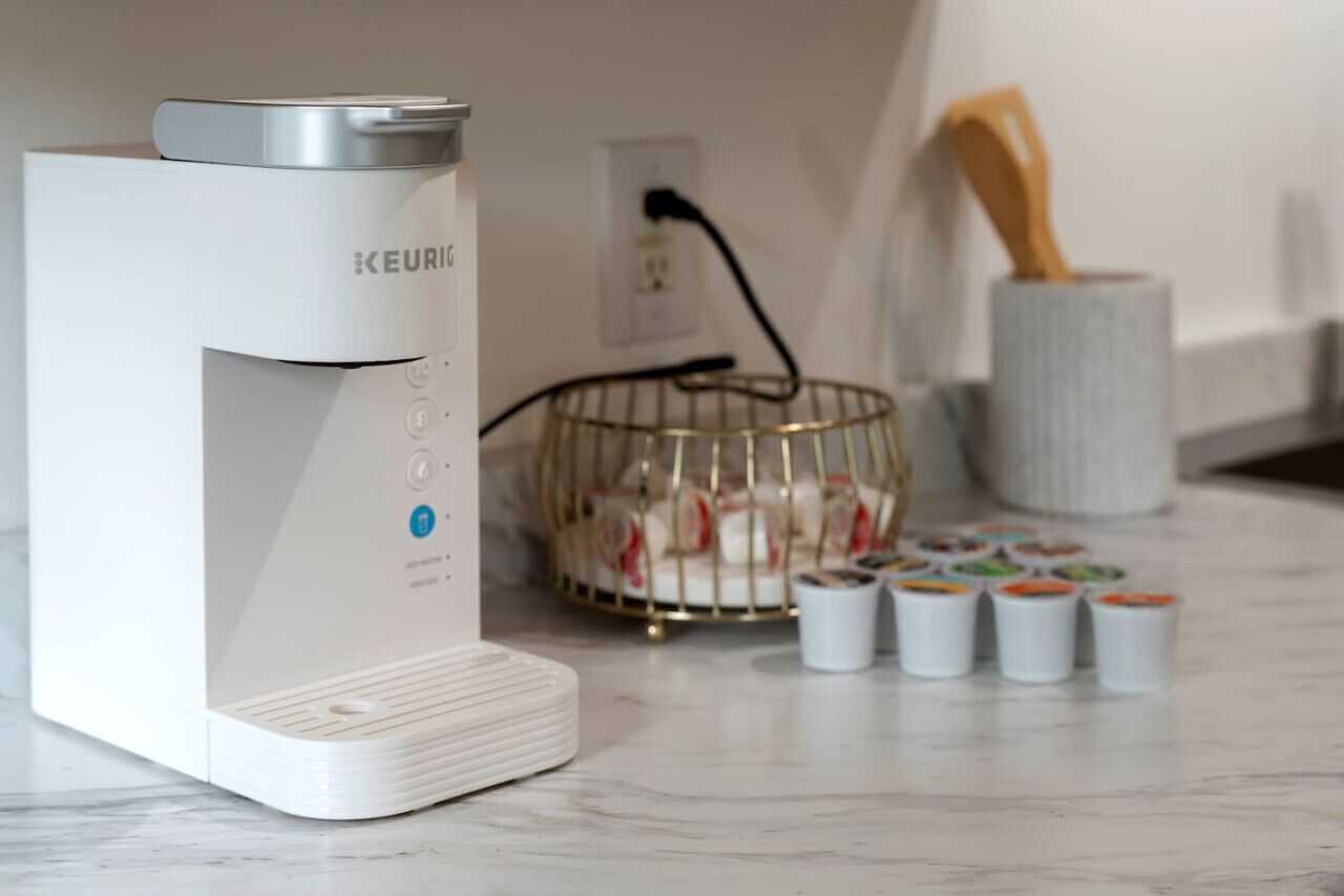 A white Keurig coffee maker on a marble countertop with a basket of coffee pods and a wooden spoon.
