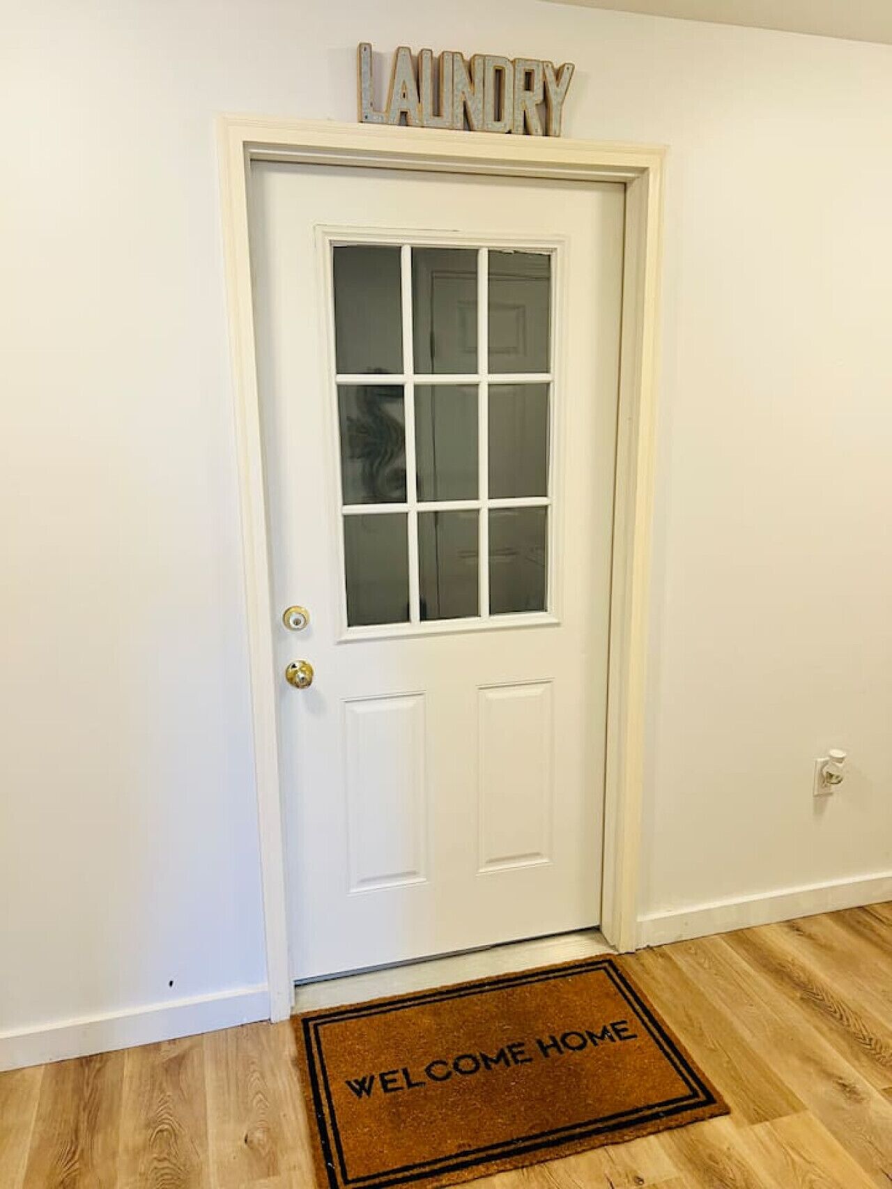 A white door with a frosted glass panel, a doormat with "WELCOME HOME" text, and a sign above the door reading "LAUNDRY".