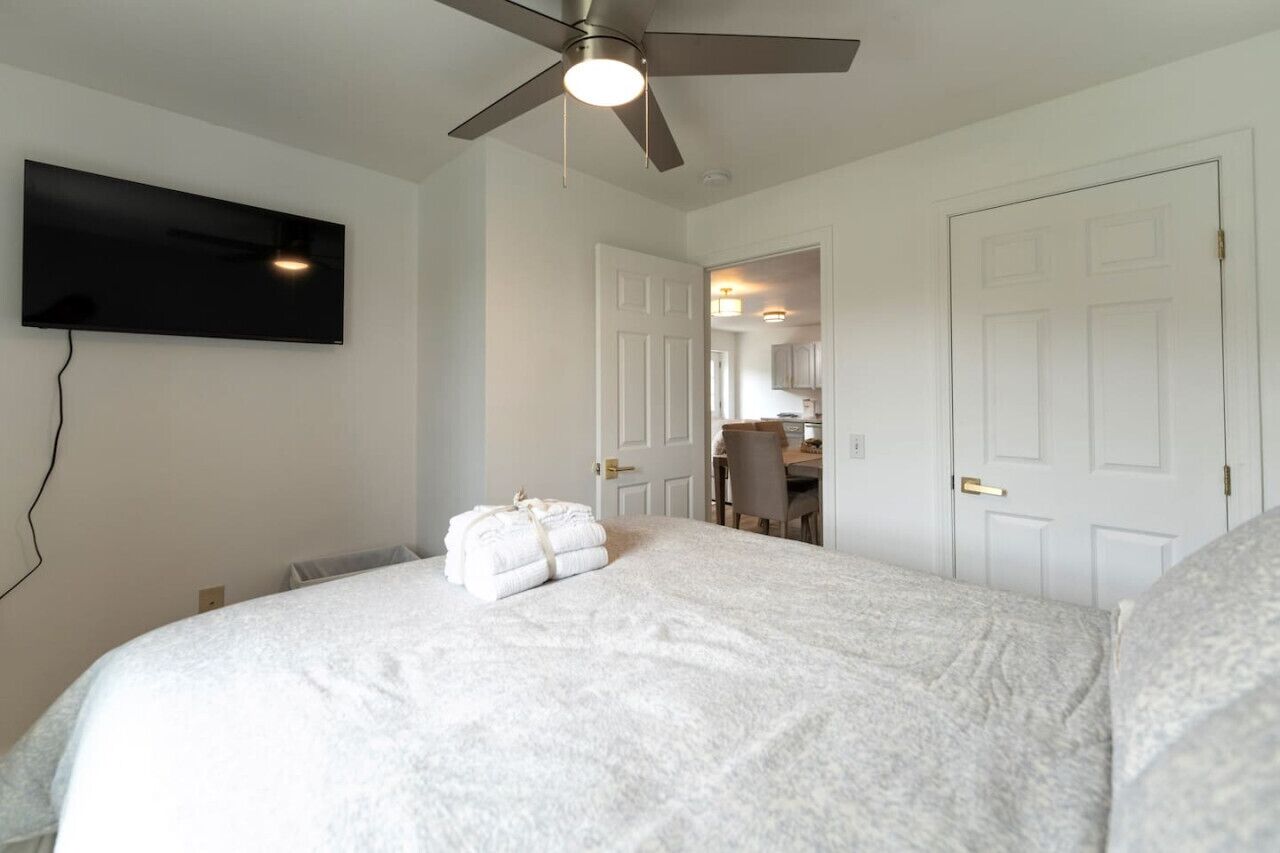 A bedroom with a bed, a ceiling fan, a TV mounted on the wall, and a door leading to a bathroom.