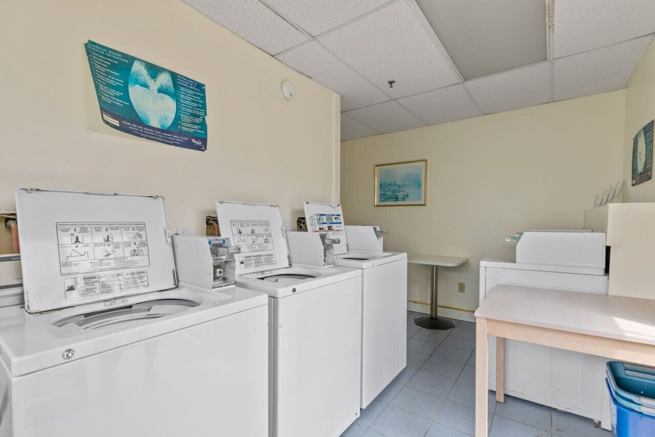 Une buanderie équipée de quatre machines à laver et de quatre sèche-linge, d’une petite table et d’une poubelle bleue.