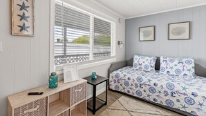 A bedroom with a bed, a window with blinds, a small table with a vase, and a wall with framed pictures. - Stylish 1BR Suite Steps to the Beach 467 (Old Orchard Beach)