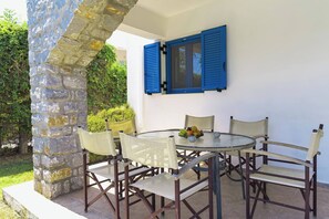 Una mesa redonda con seis sillas de mimbre, un pilar de piedra y una ventana con contraventanas azules.