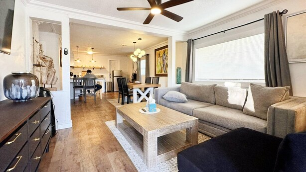 A modern living room with a wooden coffee table, a sofa, and a black ottoman. There is a ceiling fan, a dining area in the background, and a large window with curtains. - 2br-newly Renovated Full Kitchen-3 Mins Asia Dist (Oklahoma City)