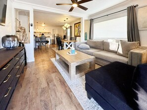 Ein modernes Wohnzimmer mit Couch, niedrigem Hocker und Couchtisch aus Holz. Ein Deckenventilator, eine Essecke im Hintergrund und ein großes Fenster mit Vorhängen sind ebenfalls zu sehen.