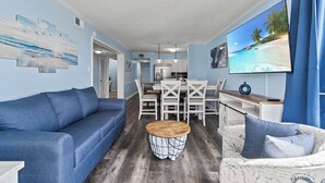 Una sala de estar con un sofá azul, una mesa de centro redonda y un televisor que muestra una escena de playa.