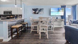 Una cocina moderna con una isla de mármol, taburetes de bar y un comedor con un televisor grande que muestra una escena de playa.
