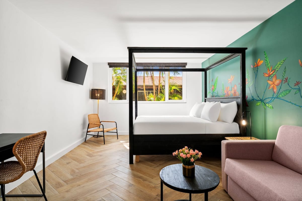 A modern room with a four-poster bed, a desk, a chair, a sofa, a round table with flowers, and a wall-mounted TV.