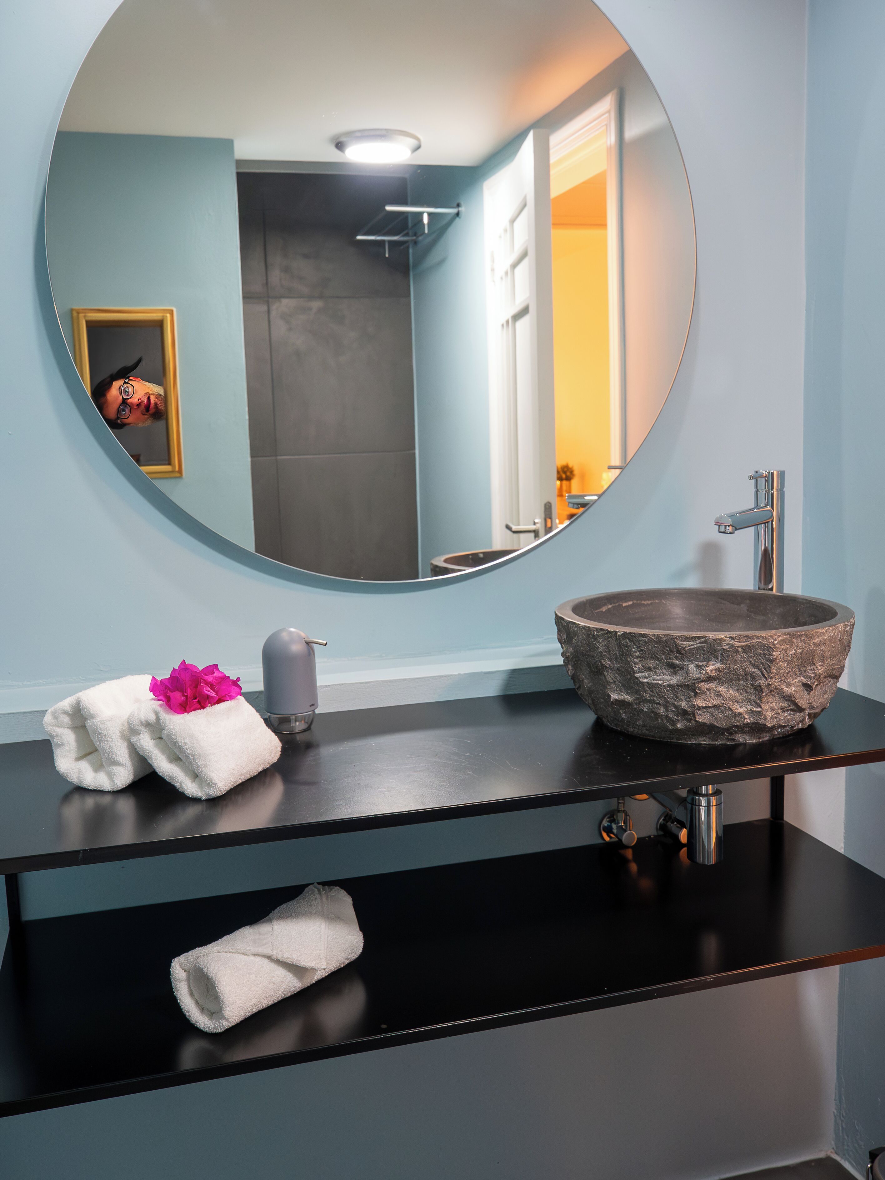 A bathroom with a round mirror, a stone sink, and a towel with a pink flower.