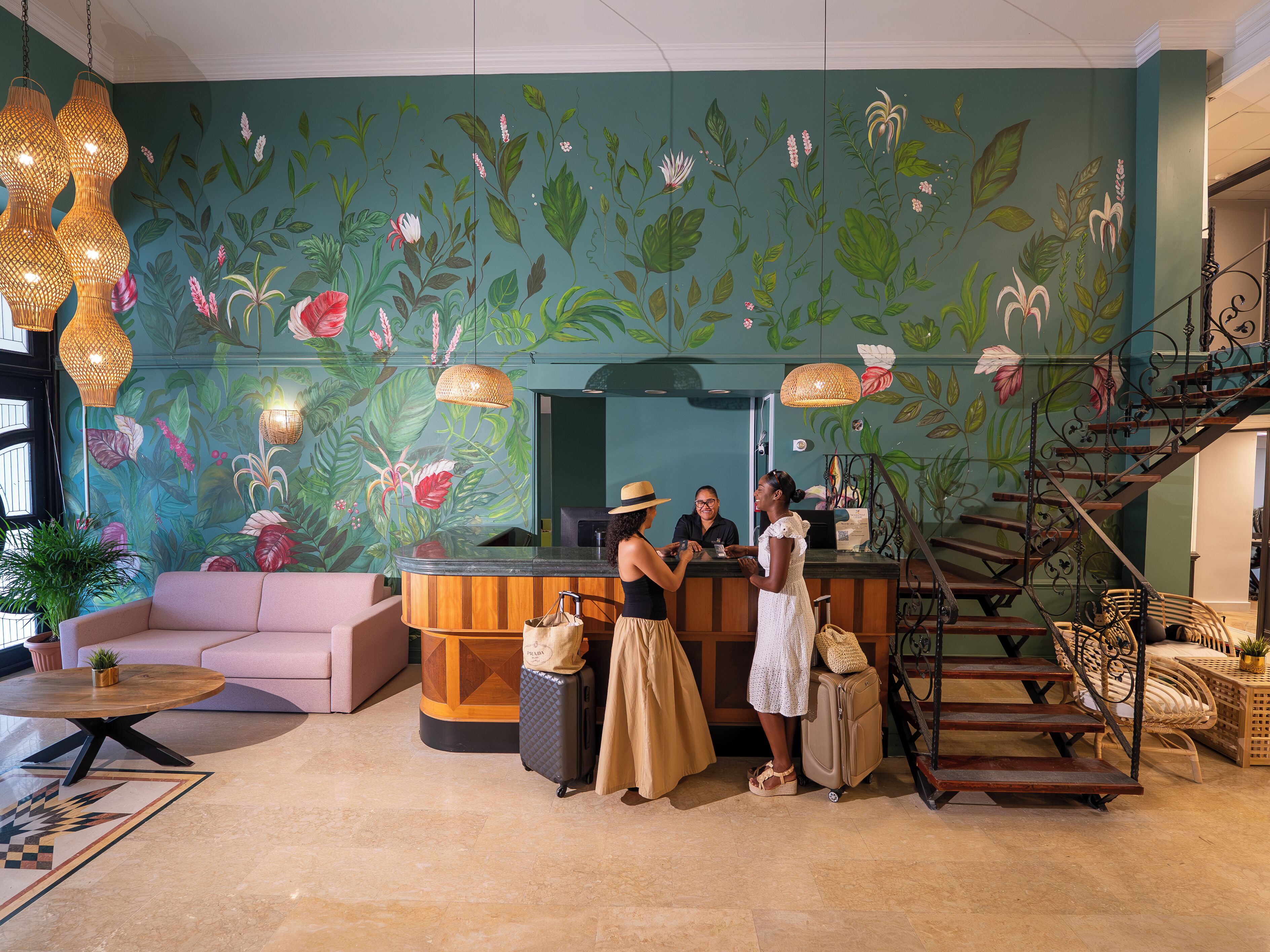 Un espace d’accueil agrémenté d’une fresque florale, un comptoir d’accueil en bois, un escalier et un coin salon avec un canapé et une table ronde.