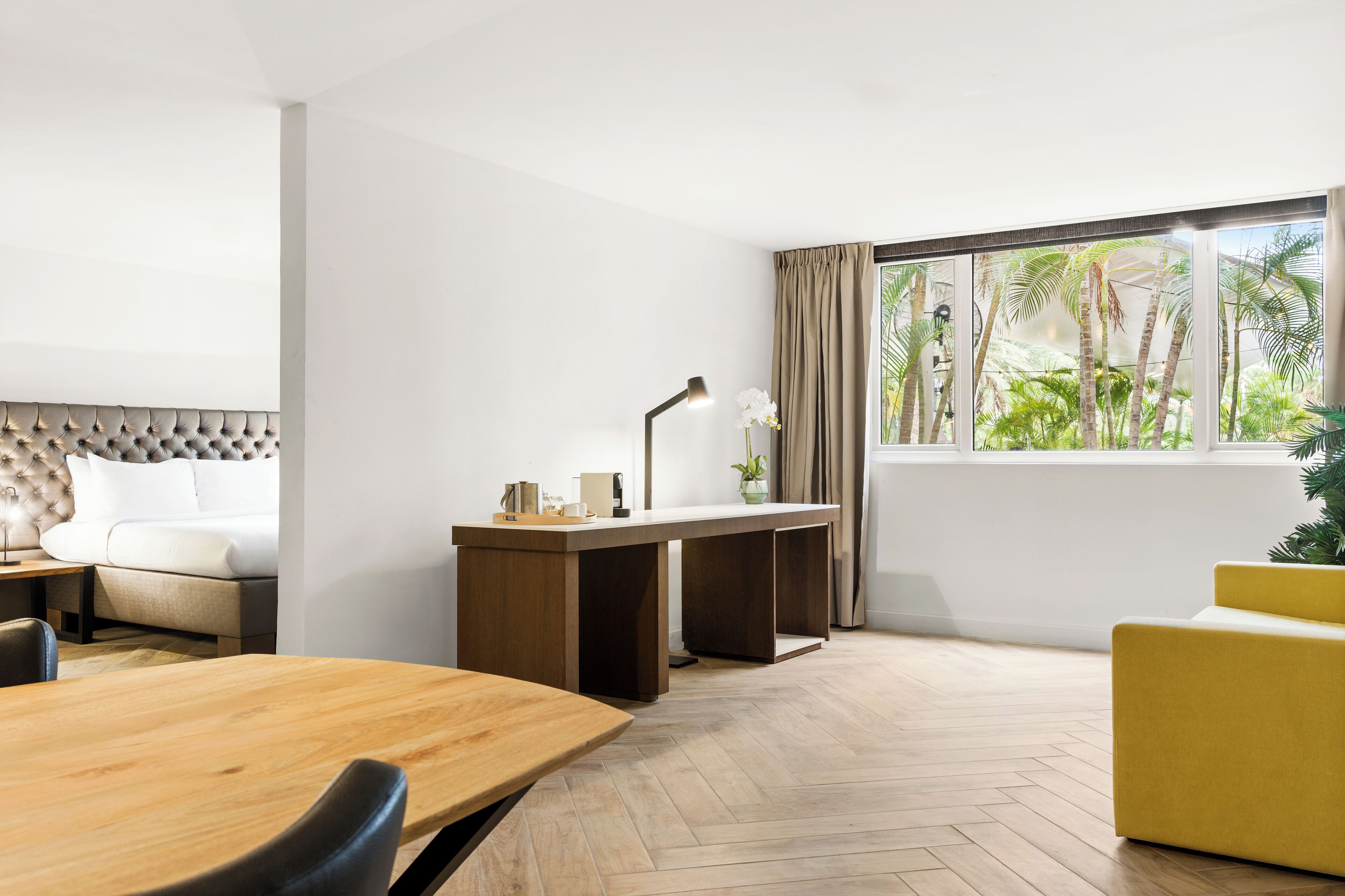Ein modernes Hotelzimmer mit einem Holztisch, einer Sitzecke, einem Schreibtisch mit Lampe und einem großen Fenster mit Blick auf Palmen.