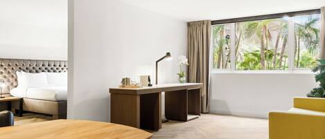 Una habitación de hotel moderna con una mesa de comedor de madera, un sofá, un escritorio con lámpara y un ventanal con vistas a palmeras.