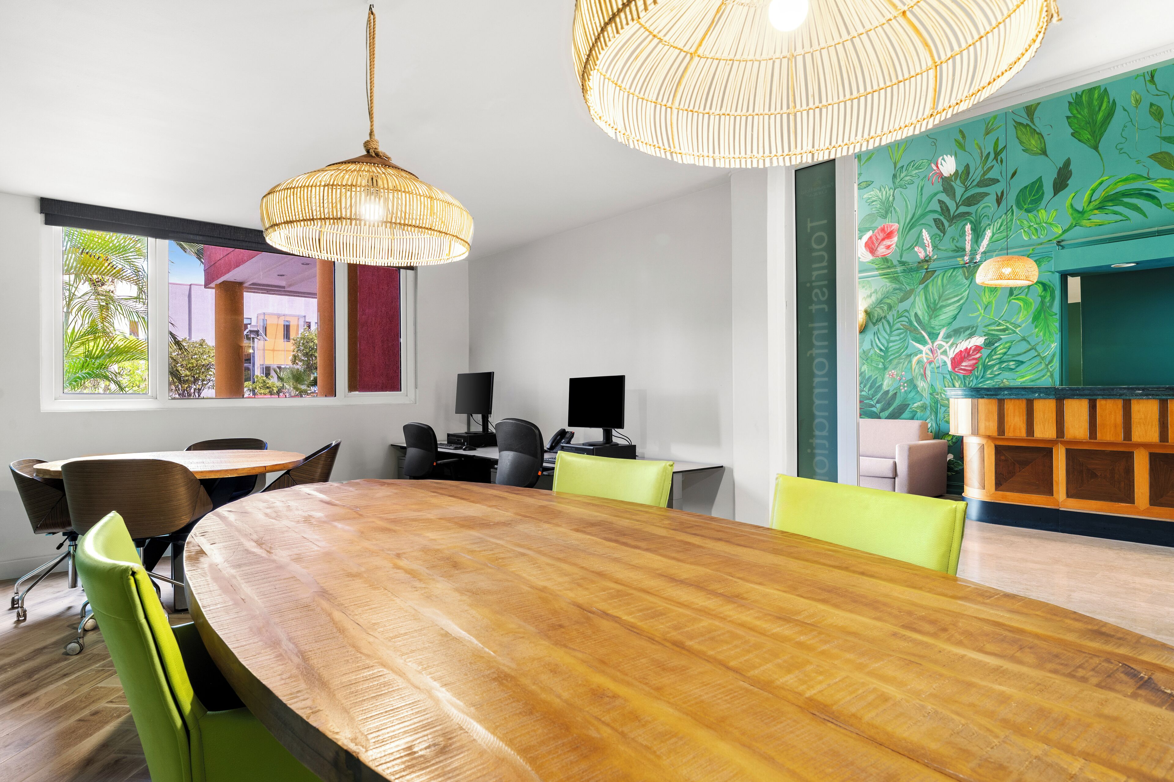 Uma sala de conferências com uma grande mesa de madeira, cadeiras verdes e duas luminárias de vime suspensas.