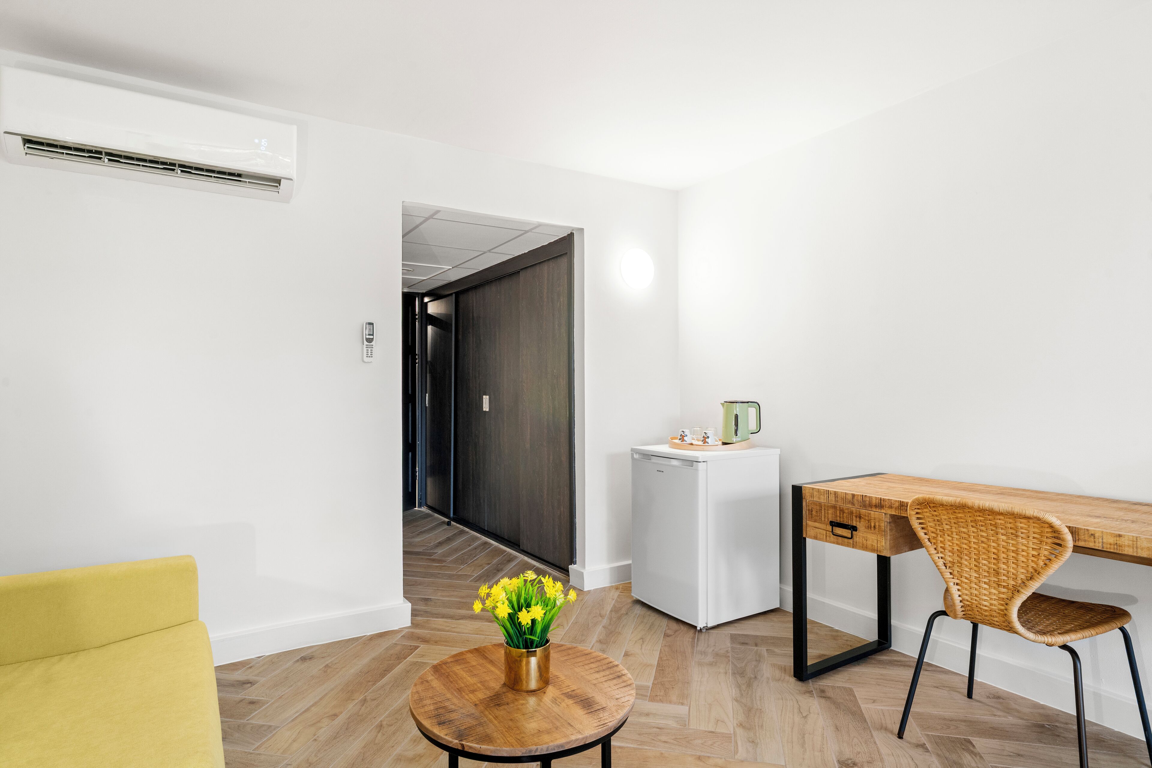 Ein modernes Zimmer mit einem gelben Sofa, einem Holztisch, einem weißen Kühlschrank und einer Vase mit gelben Blumen.
