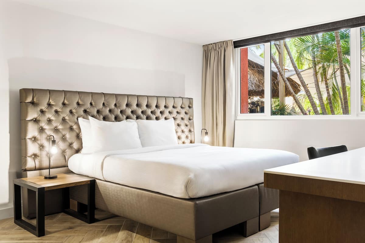 A modern hotel room with a tufted headboard, a large bed, a bedside table with a lamp, and a view of palm trees through a window.