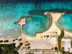 Vue aérienne d’une zone côtière avec une plage de sable, un parking et un bâtiment doté d’un toit plat.