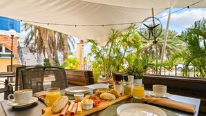 A breakfast setup with a variety of foods and drinks on a wooden board, a cup of coffee, and a plate of eggs, set in an outdoor dining area with palm trees and a view of buildings in the background. - Parasasa Hotel Curacao (Willemstad)